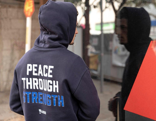 A person wearing a navy blue hoodie with 'PEACE THROUGH STRENGTH' printed on the back in white and blue, standing outdoors near a reflective glass surface.
